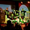 Un gruppo di persone in posa su un palco, con luci colorate e pubblico in ombra.