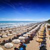 Una spiaggia affollata con file di ombrelloni bianchi e beige sotto un cielo blu.