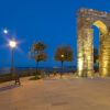Arco di pietra illuminato su piazza alberata al tramonto.