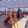 Area giochi sulla spiaggia con altalene e scivoli sotto una tettoia a righe blu.